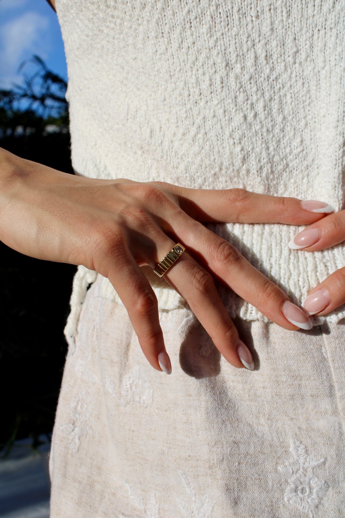 Close-up of the Vanessa ring worn on a hand, showing its ridged gold-tone band and square stone against a white outfit.