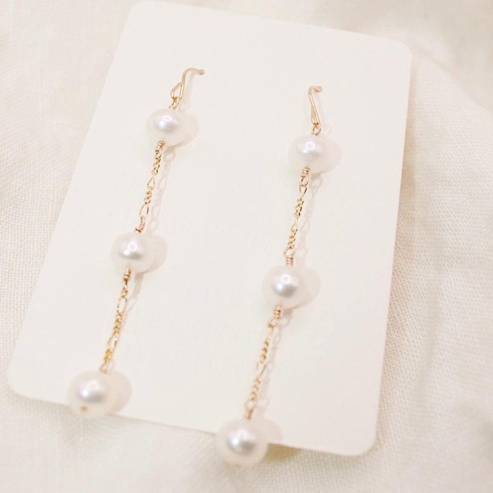 Heavenly earrings with three round white pearls spaced along each long gold-colored chain, displayed on a cream card.