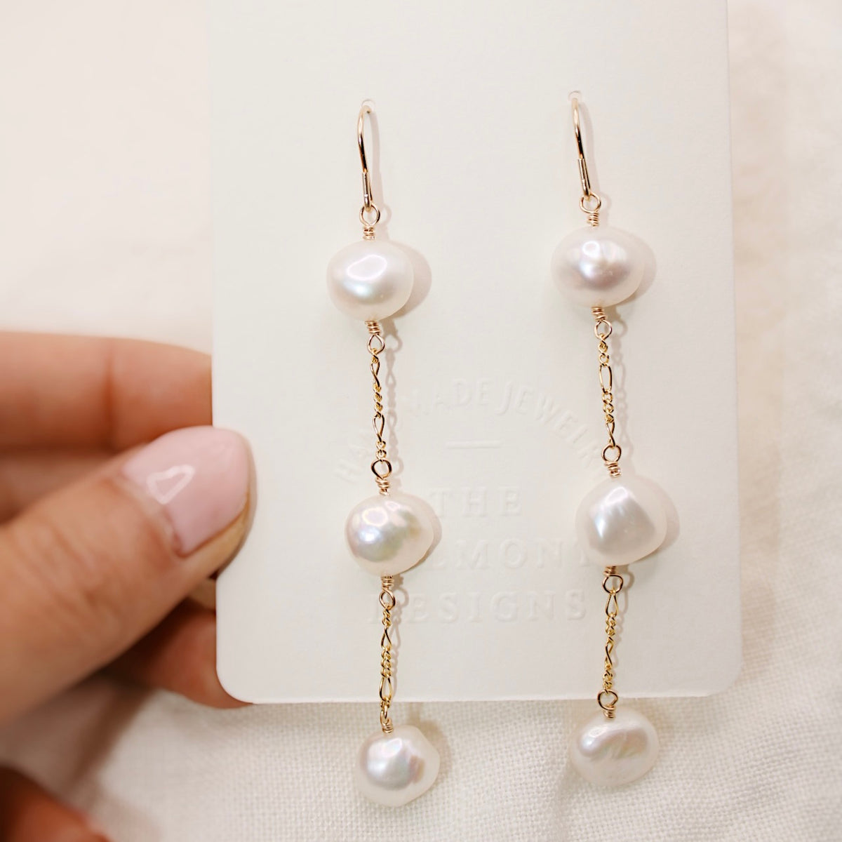 A hand holds a display card with Heavenly earrings, showing three white nugget pearls along each gold-colored chain.