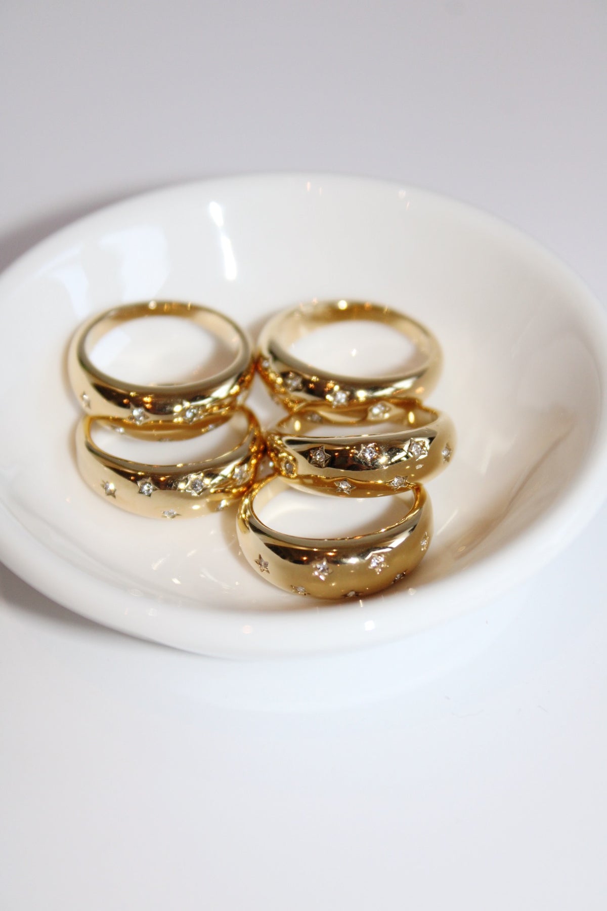Several Star Dust rings arranged in a white dish, showing their wide gold-tone bands and scattered clear stones.