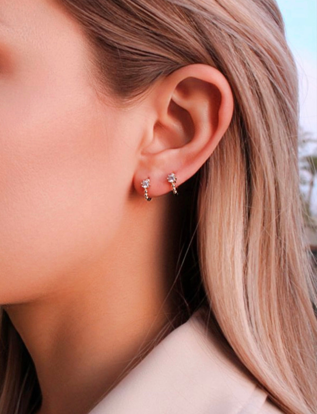 Close-up of Stacey huggie earrings worn together in two ear piercings, showing their small beaded hoops and clear stones.