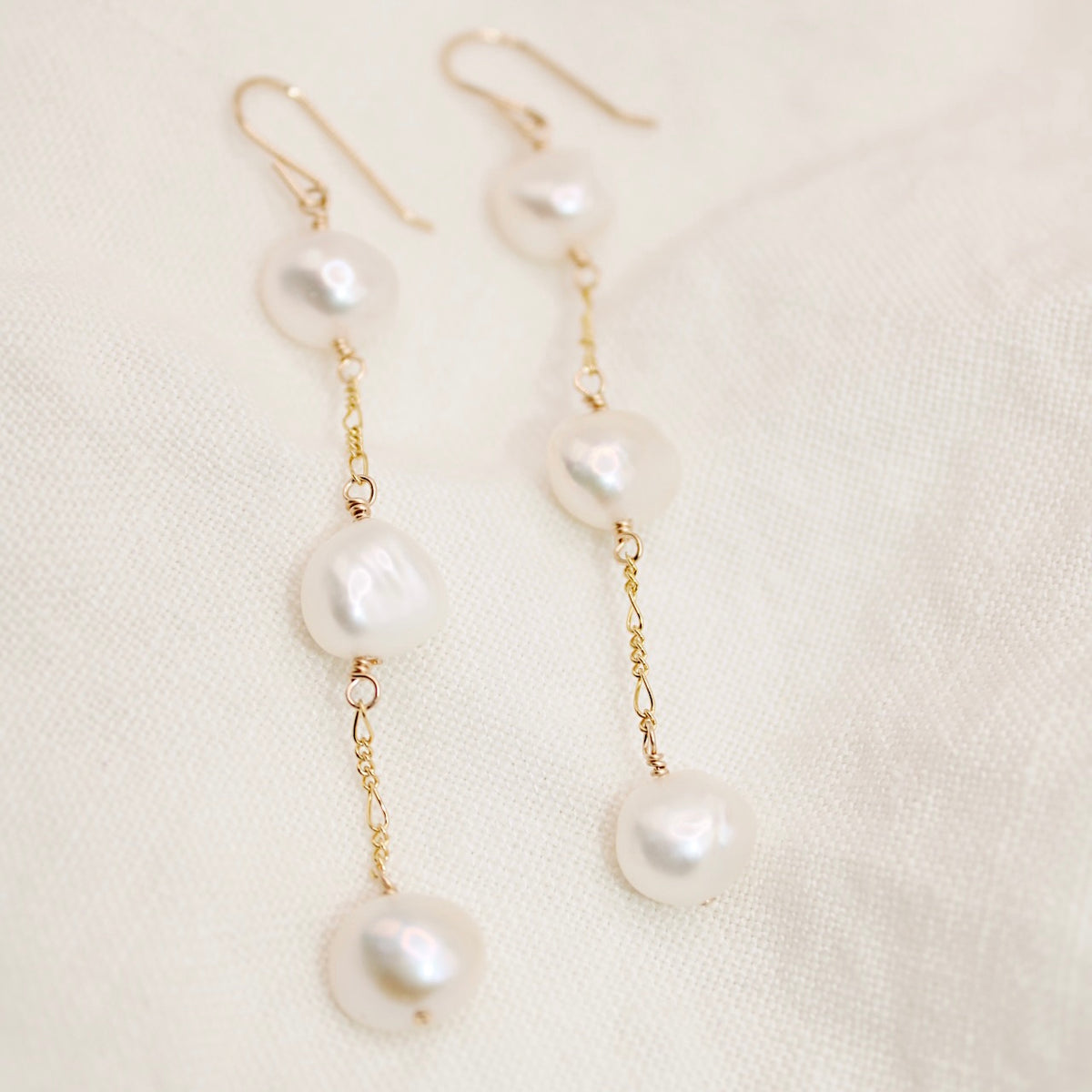 Heavenly earrings with three irregular white nugget pearls on each long gold-colored chain, resting on cream fabric.