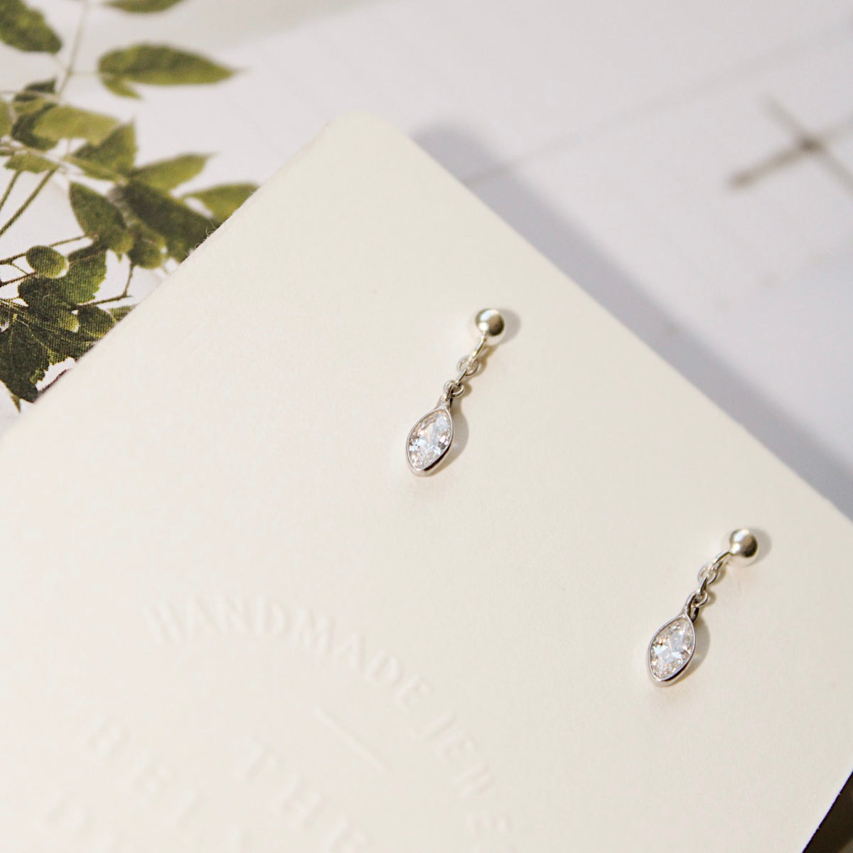 Dana silver drop earrings on an angled cream display card beside green leaves.