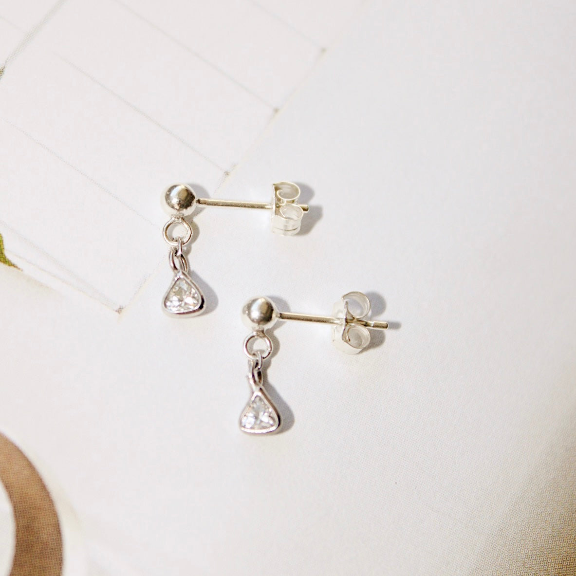Close-up of Lexy sterling silver stud earrings with dangling triangular clear stones, shown with butterfly backs on a pale surface.