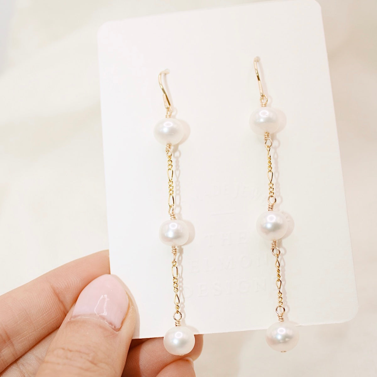 A hand holds Heavenly earrings on a cream display card, showing three round pearls on each gold-colored chain drop.
