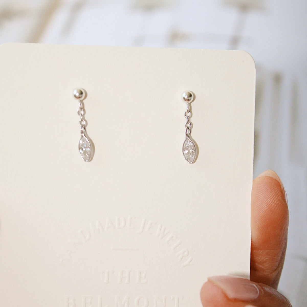 A hand holds a cream display card with Dana earrings, showing their short silver chains and clear marquise charms.