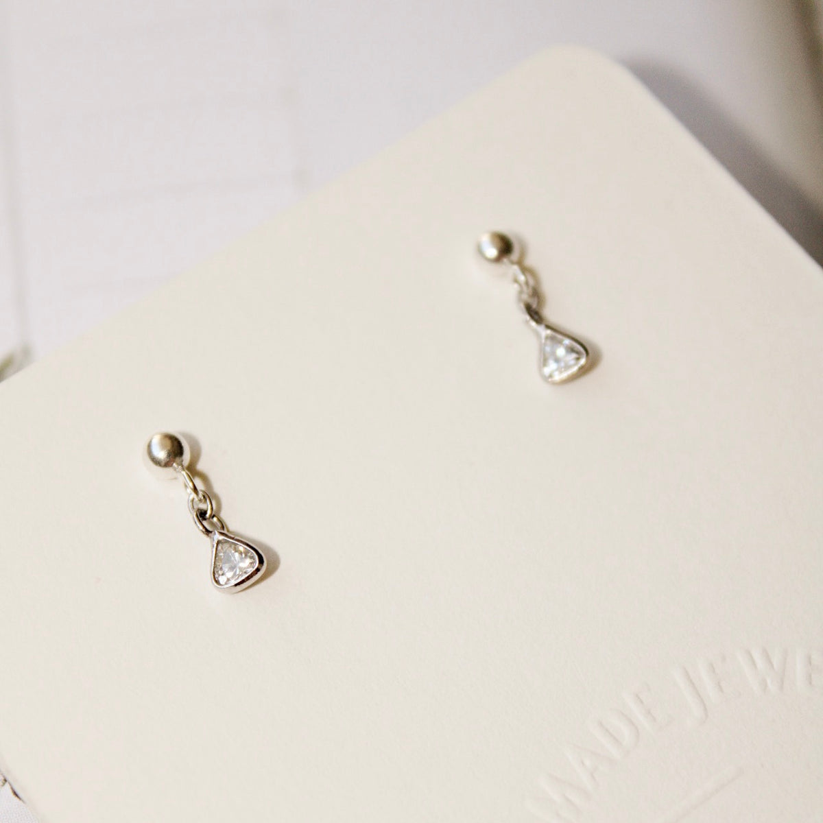 Lexy earrings displayed on a cream jewelry card, showing round silver studs and delicate triangular clear-stone drops.